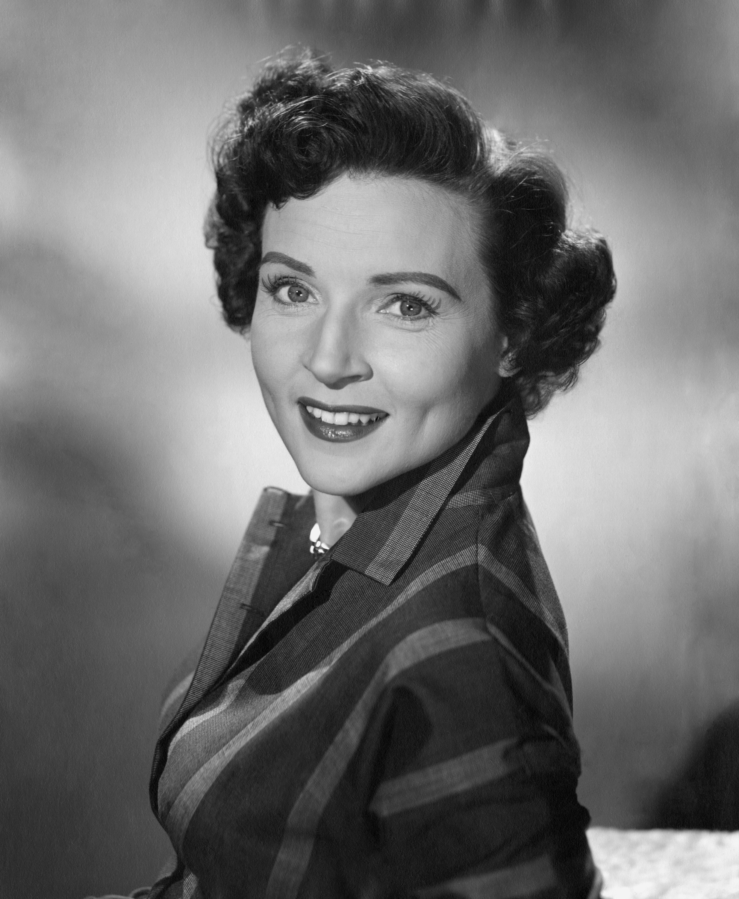 Betty White in a classic portrait, wearing a plaid jacket and smiling at the camera