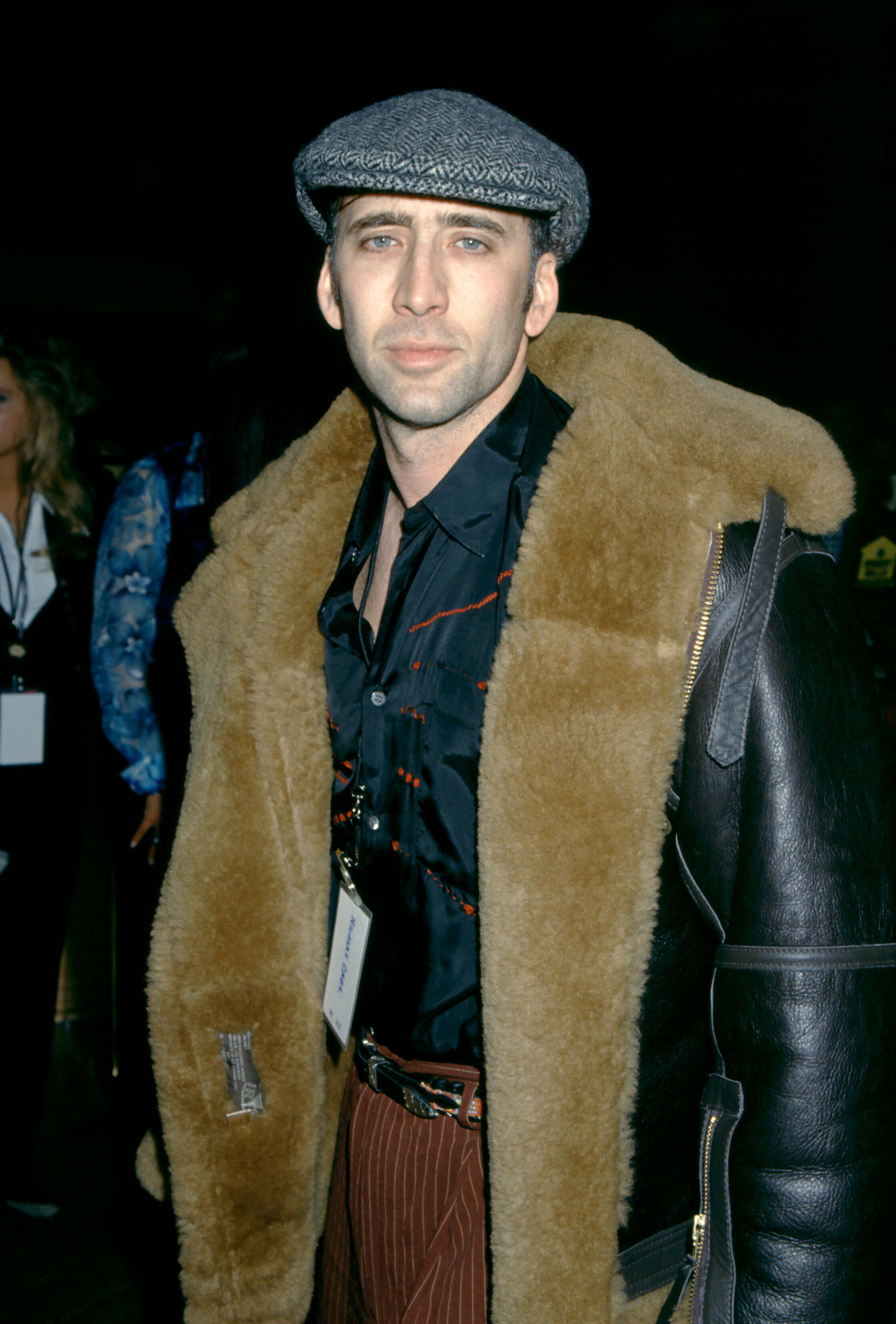 Nicolas Cage poses in a stylish outfit featuring a large fur-lined jacket and a flat cap, with people in the background at an event