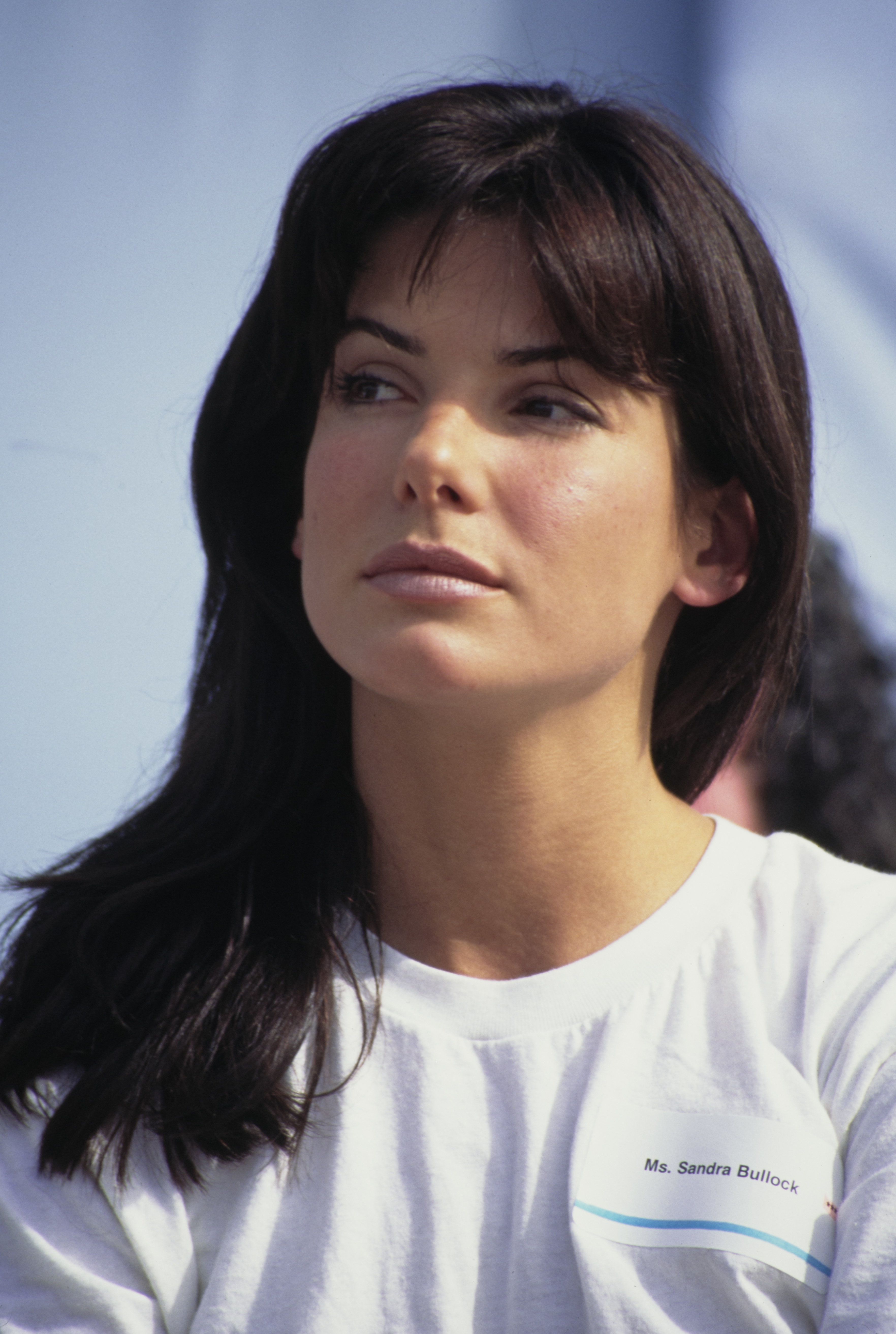 Sandra Bullock, wearing a simple white t-shirt with her name tag, looks to the side in a candid photo