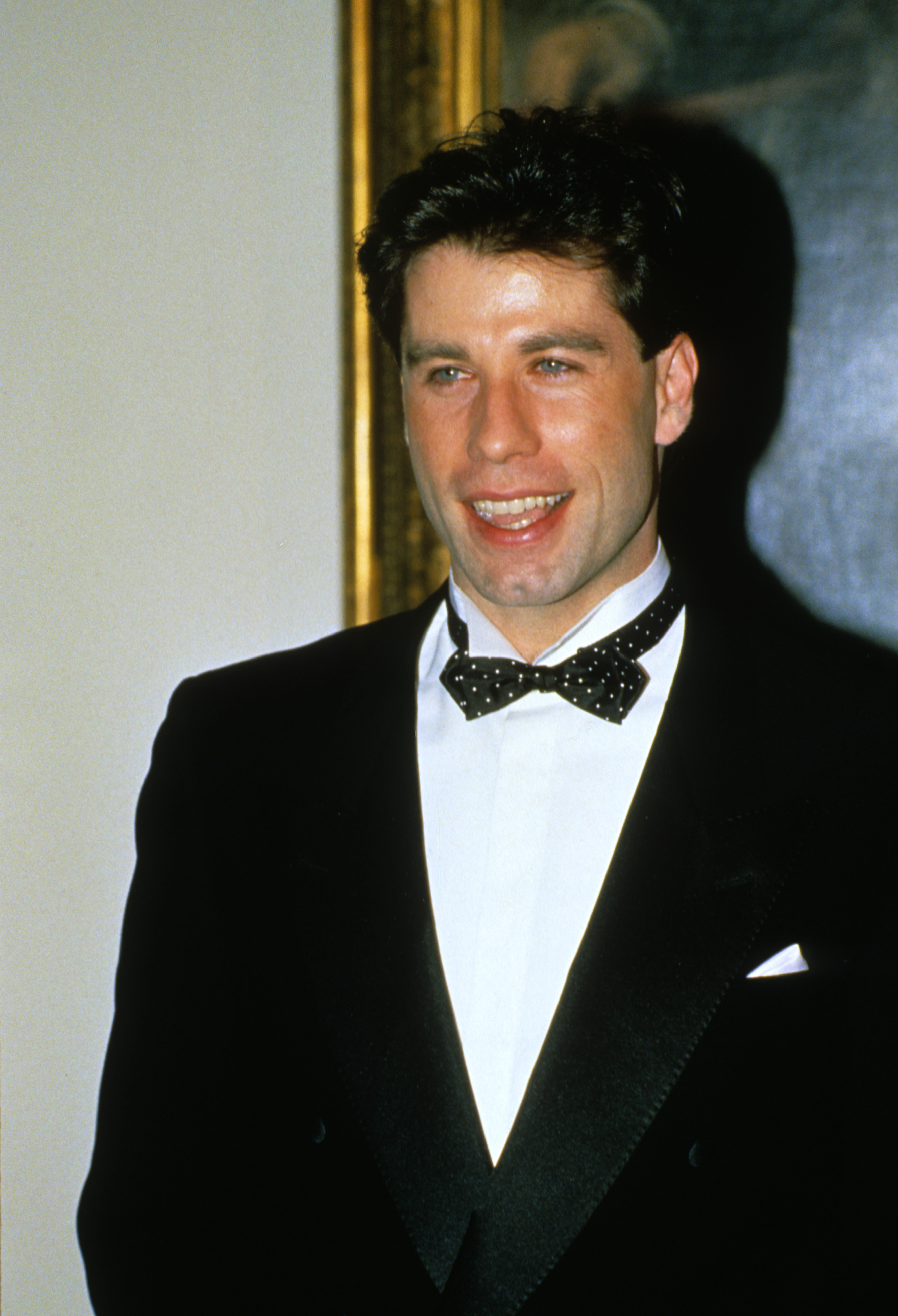 John Travolta in a black tuxedo and polka dot bow tie, smiling at an event