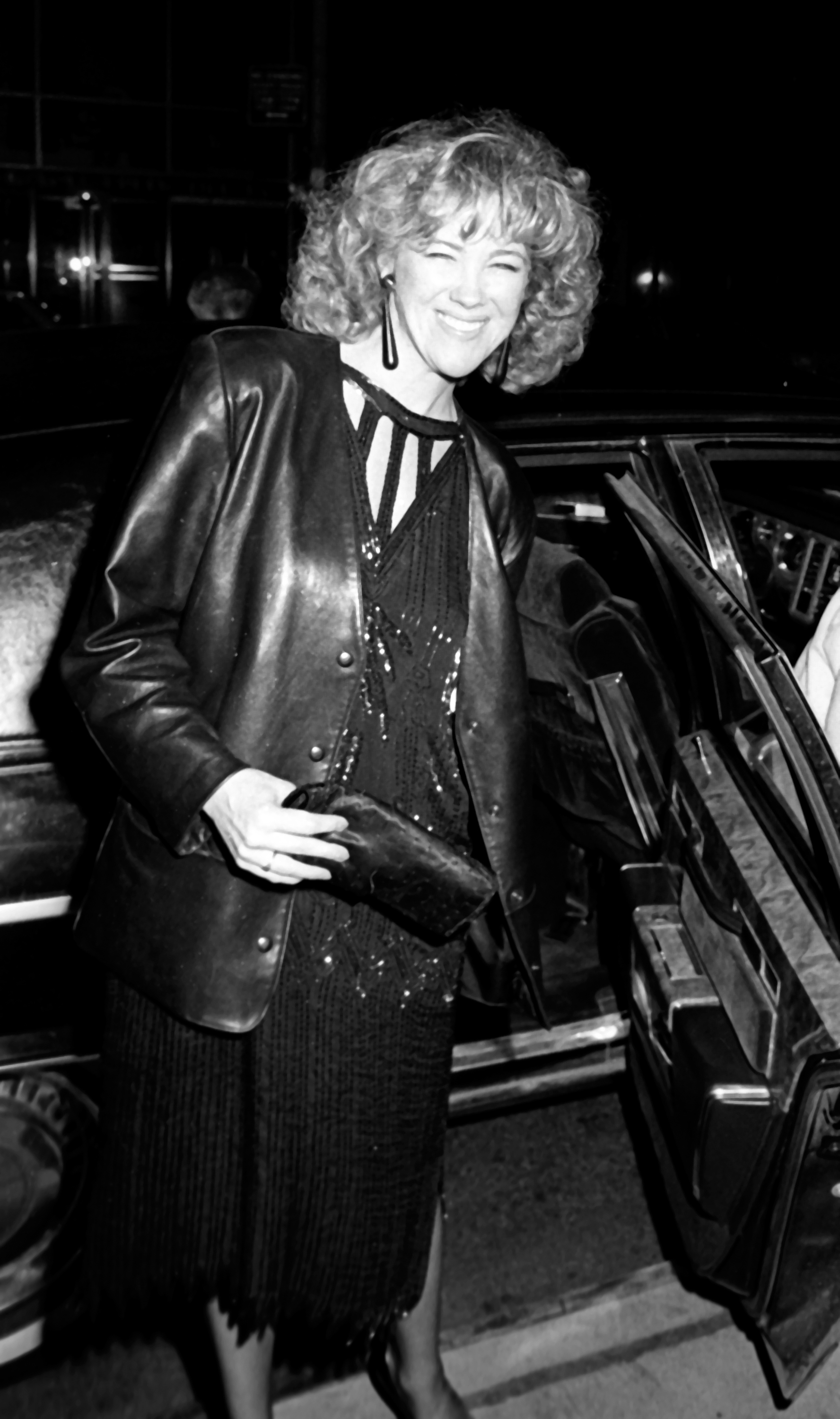 A woman with curly hair, wearing an elegant, sequined black dress and a leather jacket, steps out of a car at a nighttime event