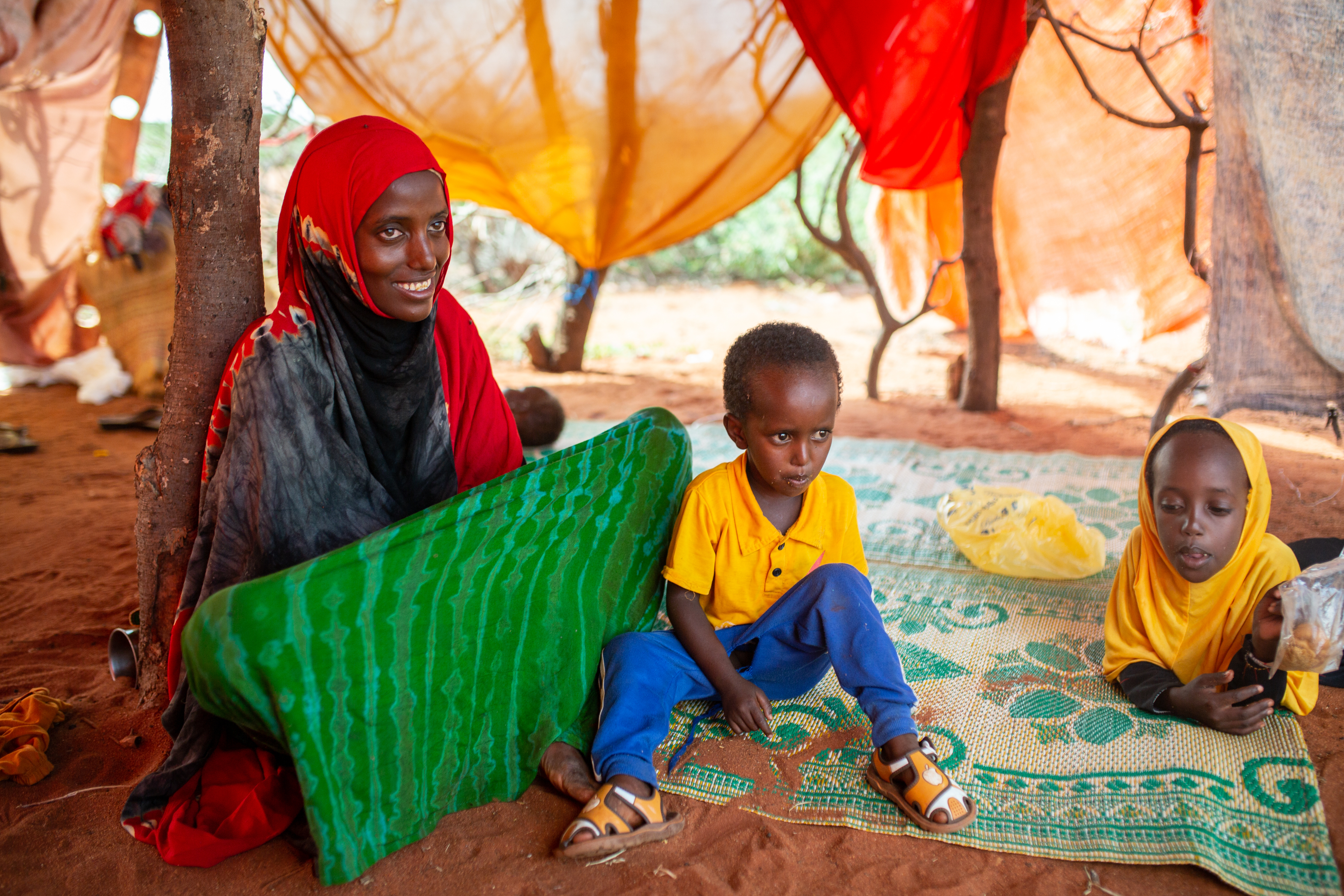 Three people, a woman and two children, sit on mats inside a tentlike structure; the woman is draped in a patterned fabric, and the children are seated nearby