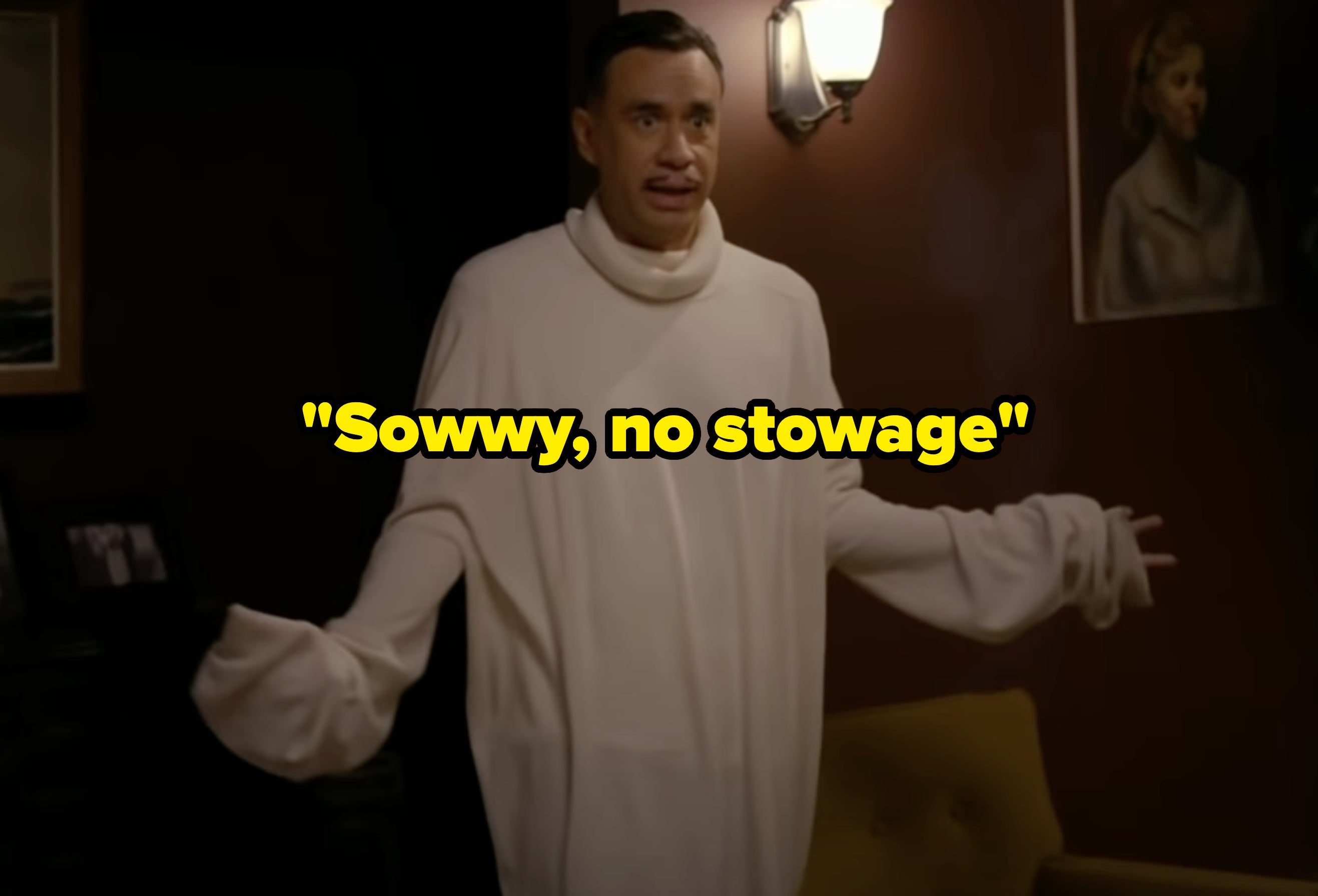 Fred Armisen stands indoors, wearing an oversized turtleneck sweater with extended sleeves, gesturing with both hands