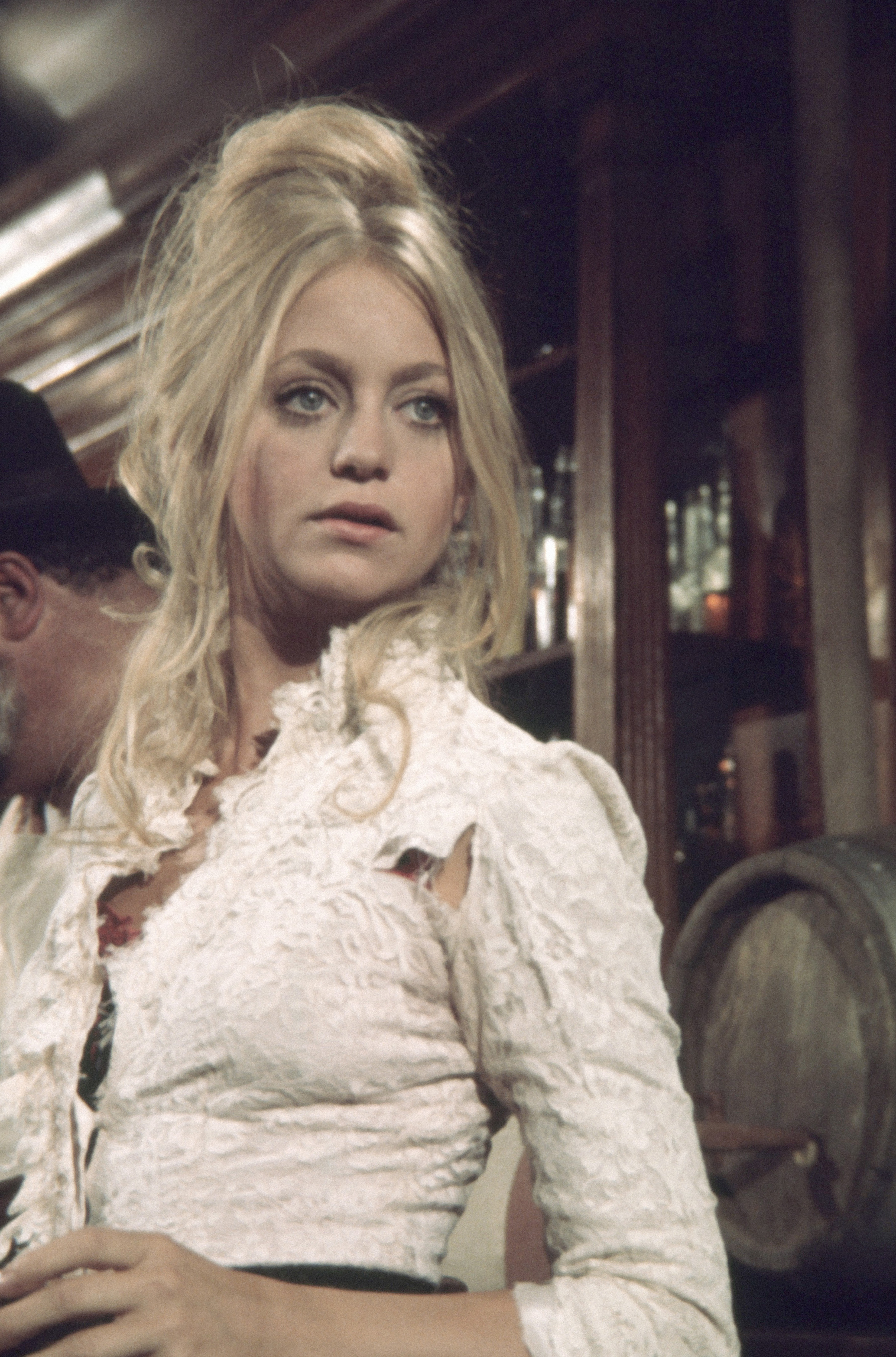 Young woman with long, wavy hair, wearing a lacy, Victorian-style blouse, standing in what looks like an old-fashioned bar