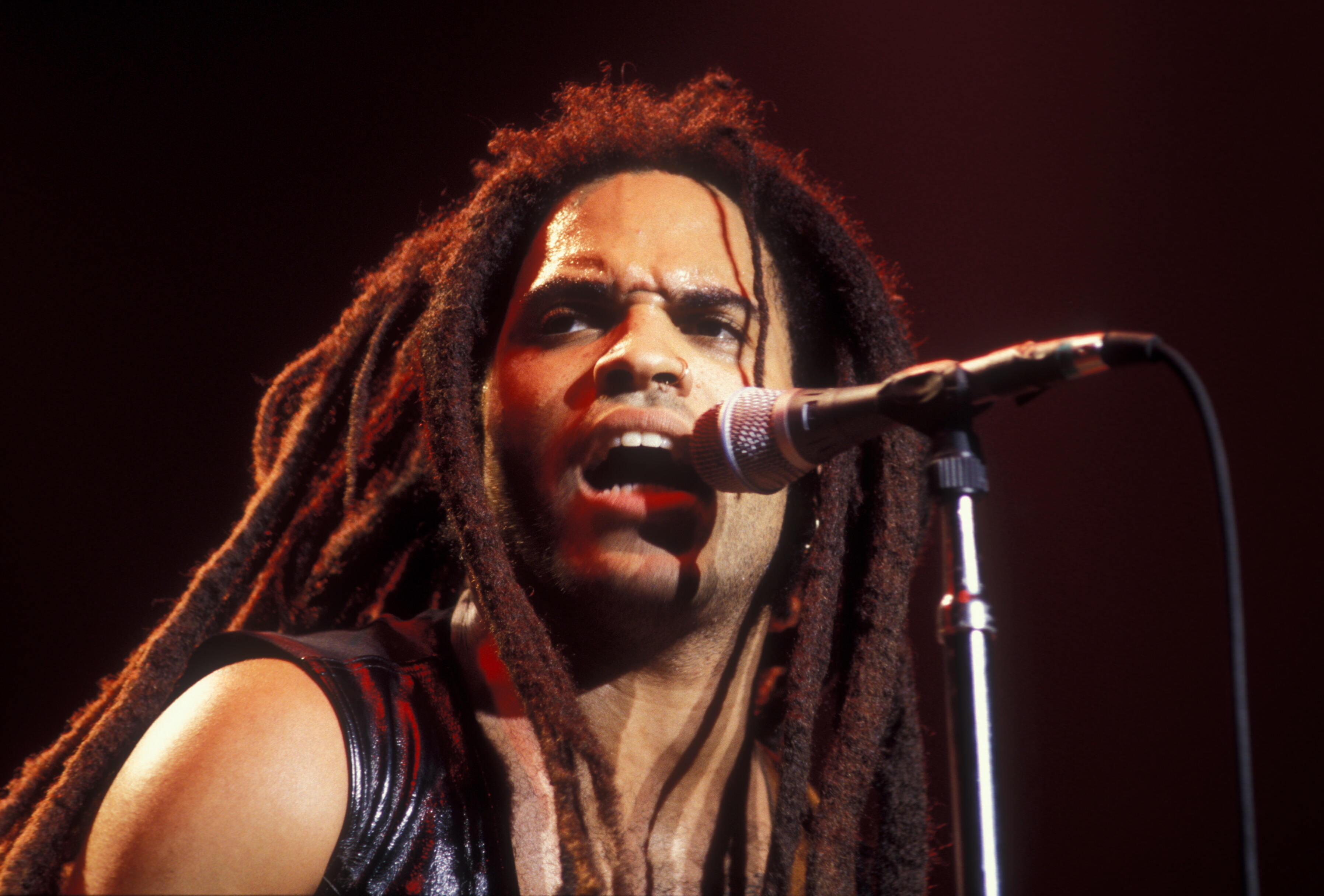 Lenny Kravitz performs live on stage, passionately singing into a microphone. He wears a leather outfit and has long dreadlocks