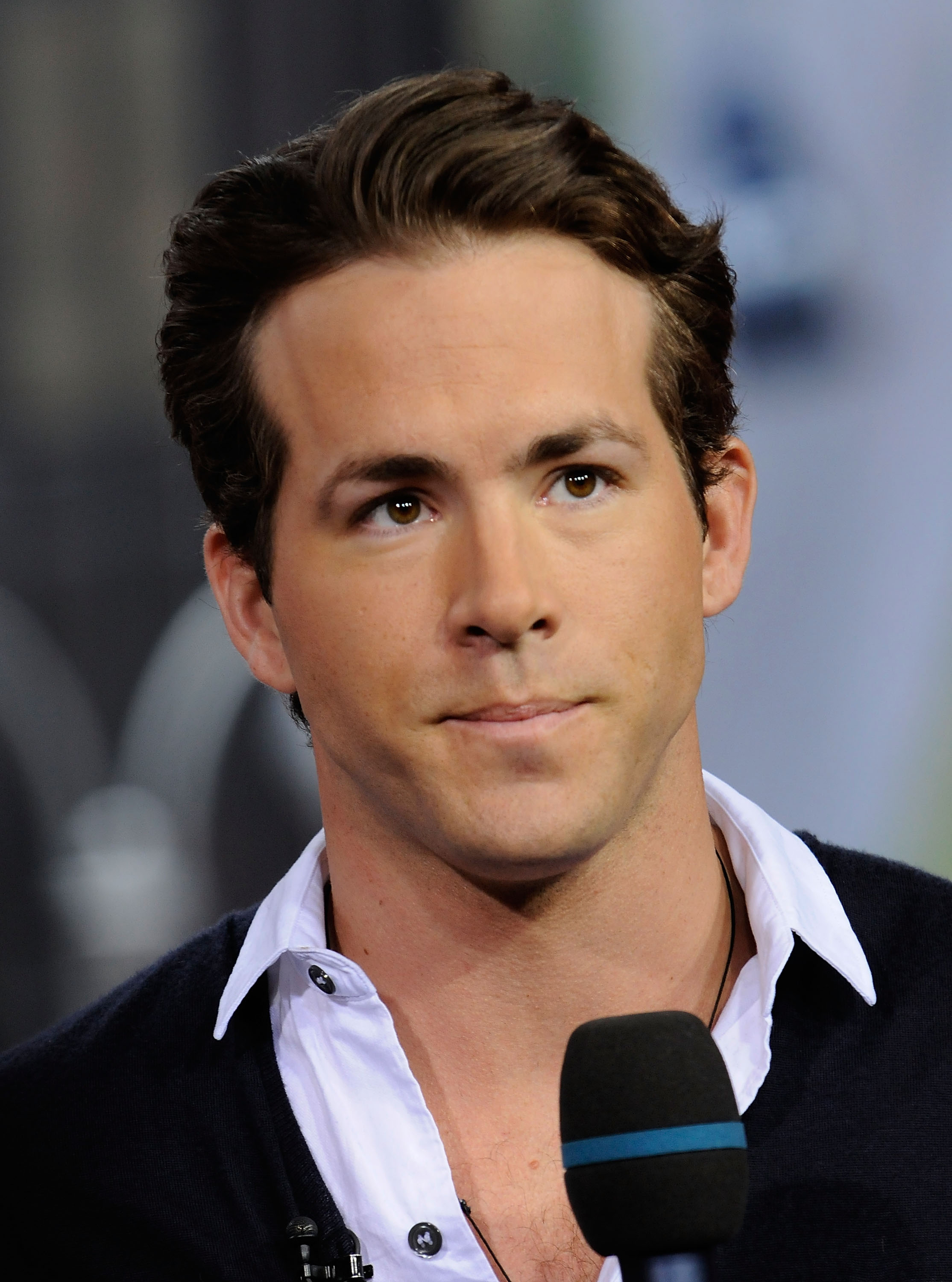 Ryan Reynolds wearing a white shirt and dark sweater, speaking into a microphone