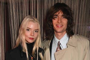Anya Taylor-Joy and Malcolm McRae posing together, with Anya wearing a black turtleneck and Malcolm in a beige trench coat