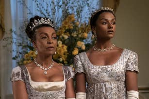 Adjoa Andoh and Simone Ashley wearing elegant gowns and tiaras, standing indoors with floral arrangements in the background