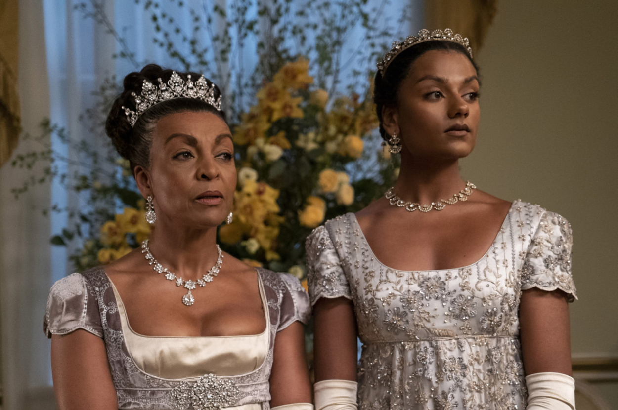 Adjoa Andoh and Simone Ashley wearing elegant gowns and tiaras, standing indoors with floral arrangements in the background