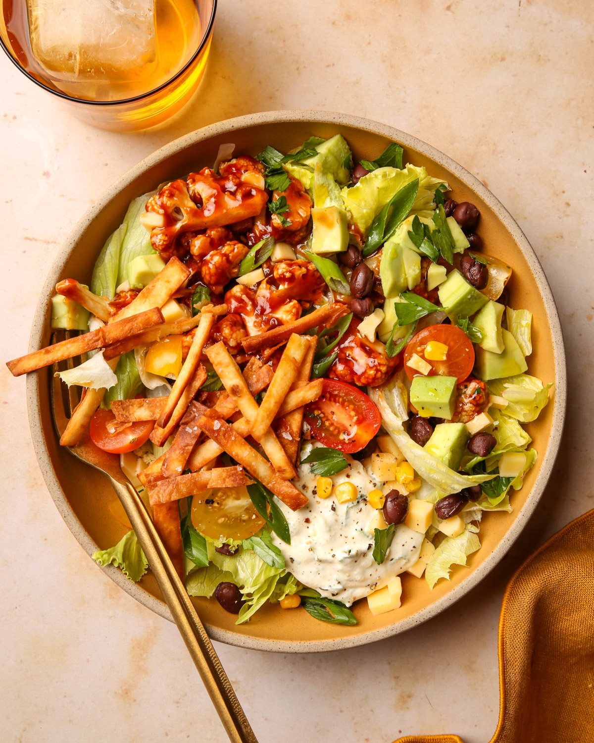 A bowl of salad with avocado, tortilla strips, cherry tomatoes, black beans, corn, and lettuce, topped with a creamy dressing and a drink with ice on the side