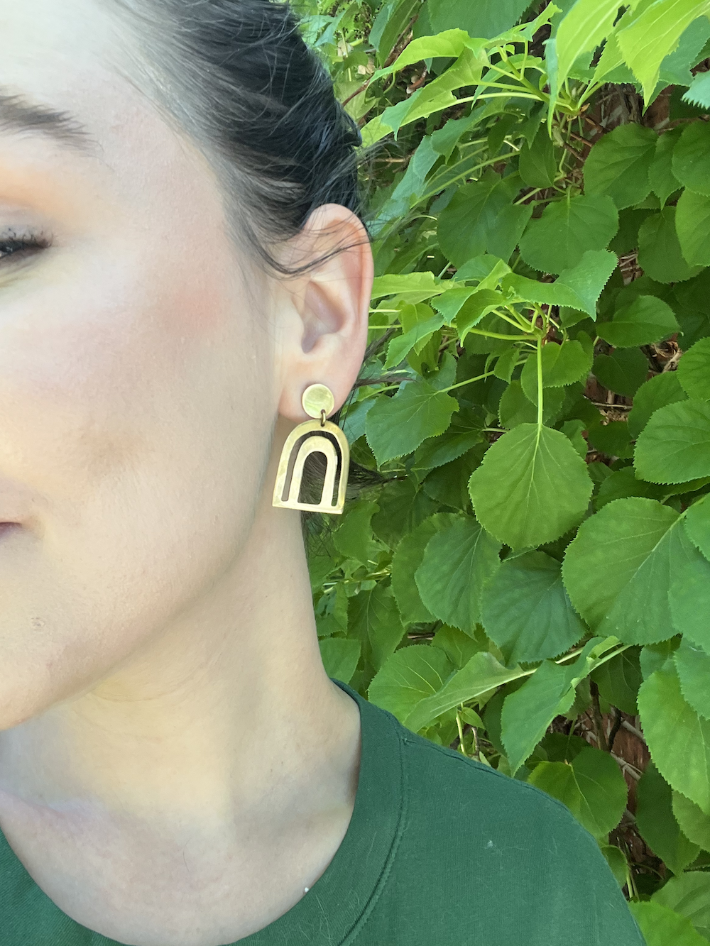 Close-up of a person wearing large, gold, arch-shaped earrings in front of green leaves