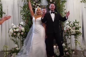 Emily and Brennan stand at their wedding ceremony, smiling with one arm raised, surrounded by greenery and flowers. Guests applaud. Caption reads "[guests cheering]"