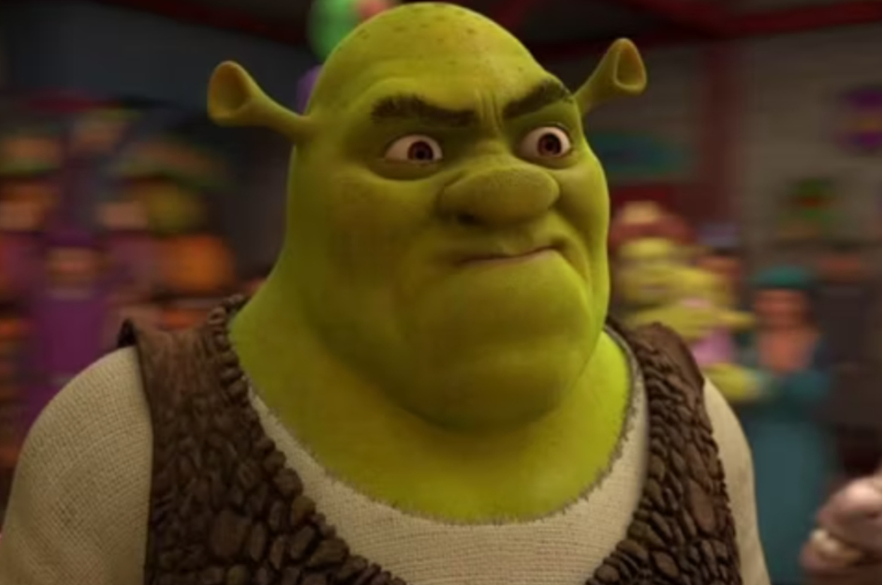 Shrek stands with a serious expression at a crowded event with blurred characters in the background