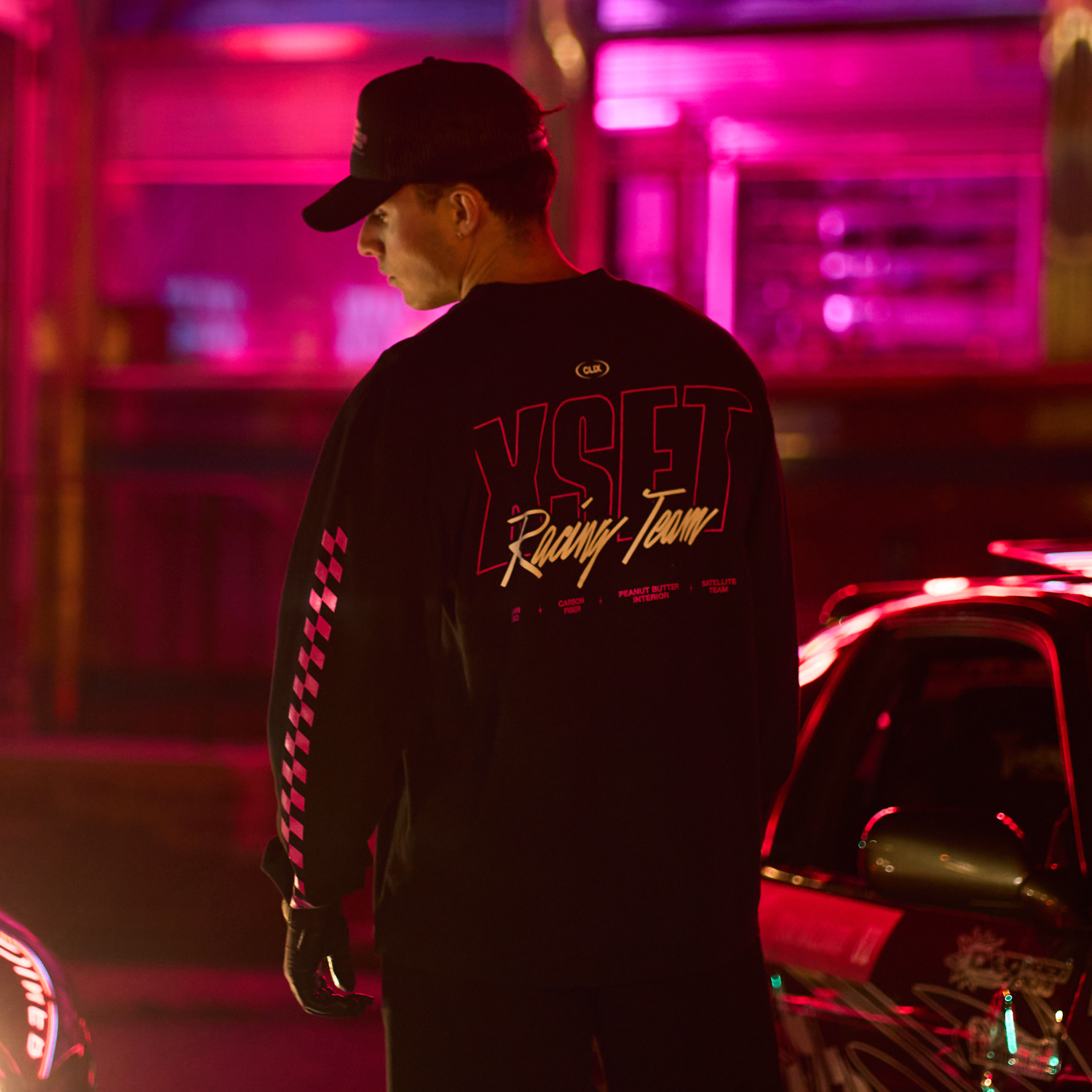 A man in a cap and YSET Racing Team jacket stands near a brightly lit car, with neon lights in the background