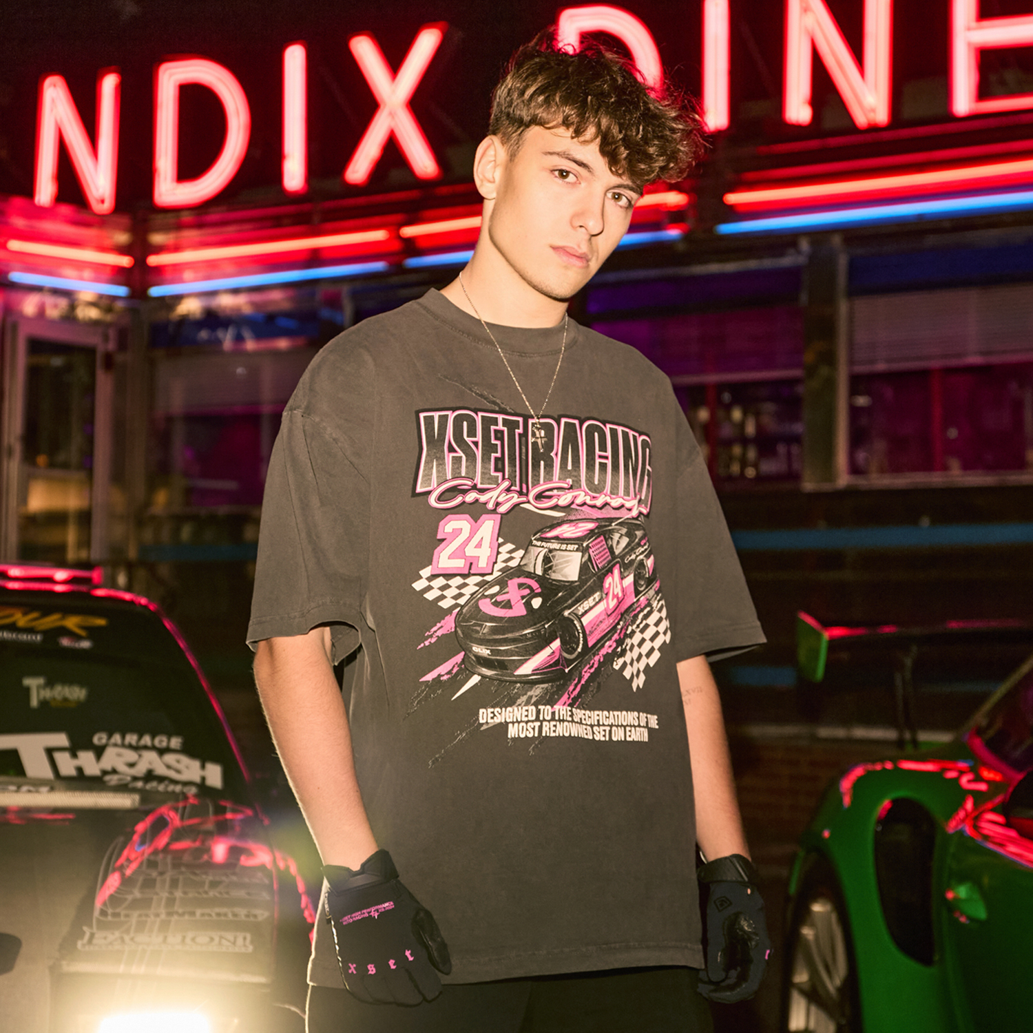 A young man stands in front of a neon-lit diner, wearing a graphic racing shirt and fingerless gloves