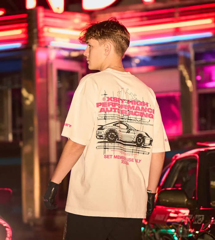 A person stands with their back turned, wearing a casual T-shirt featuring a car design and text: &quot;6-speed high performance auto racing&quot; and &quot;Set Members Only&quot;