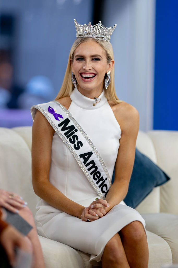 Miss America sits on a couch wearing a white sleeveless dress, her crown, and a sash. She is smiling brightly