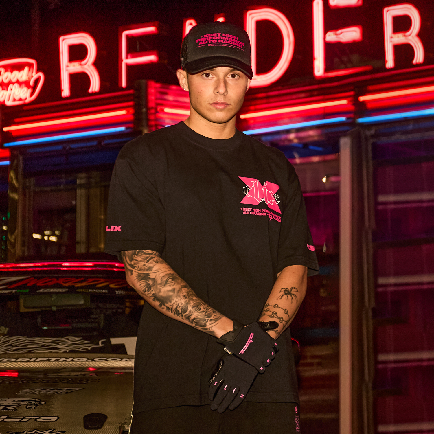 Person wearing black logo t-shirt, matching cap, and gloves, posing with tattooed arms crossed in front of neon lights