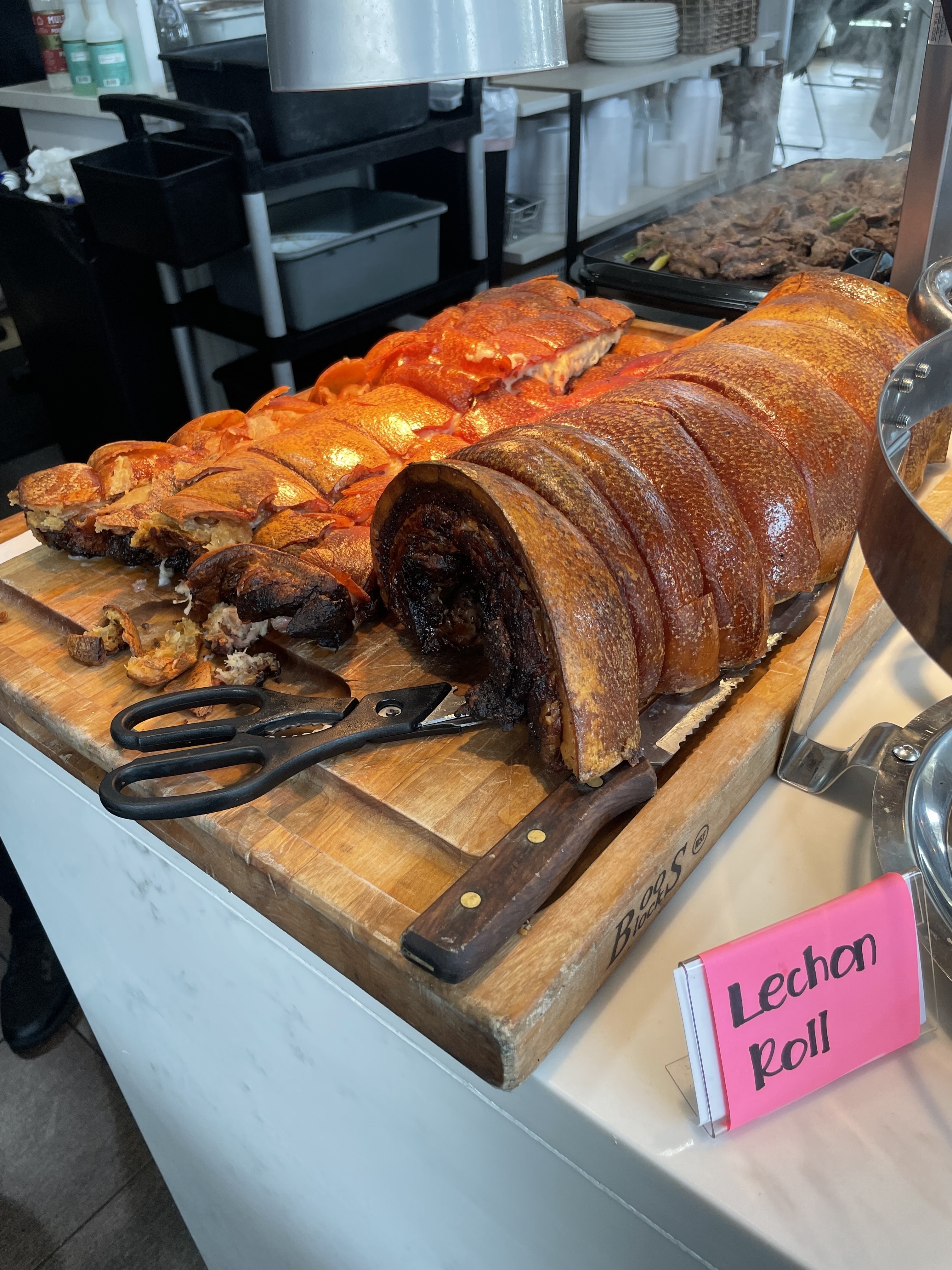 A display of Lechon Roll, a Filipino roasted pork dish, on a wooden cutting board. A sharp knife and scissors are placed beside it for serving