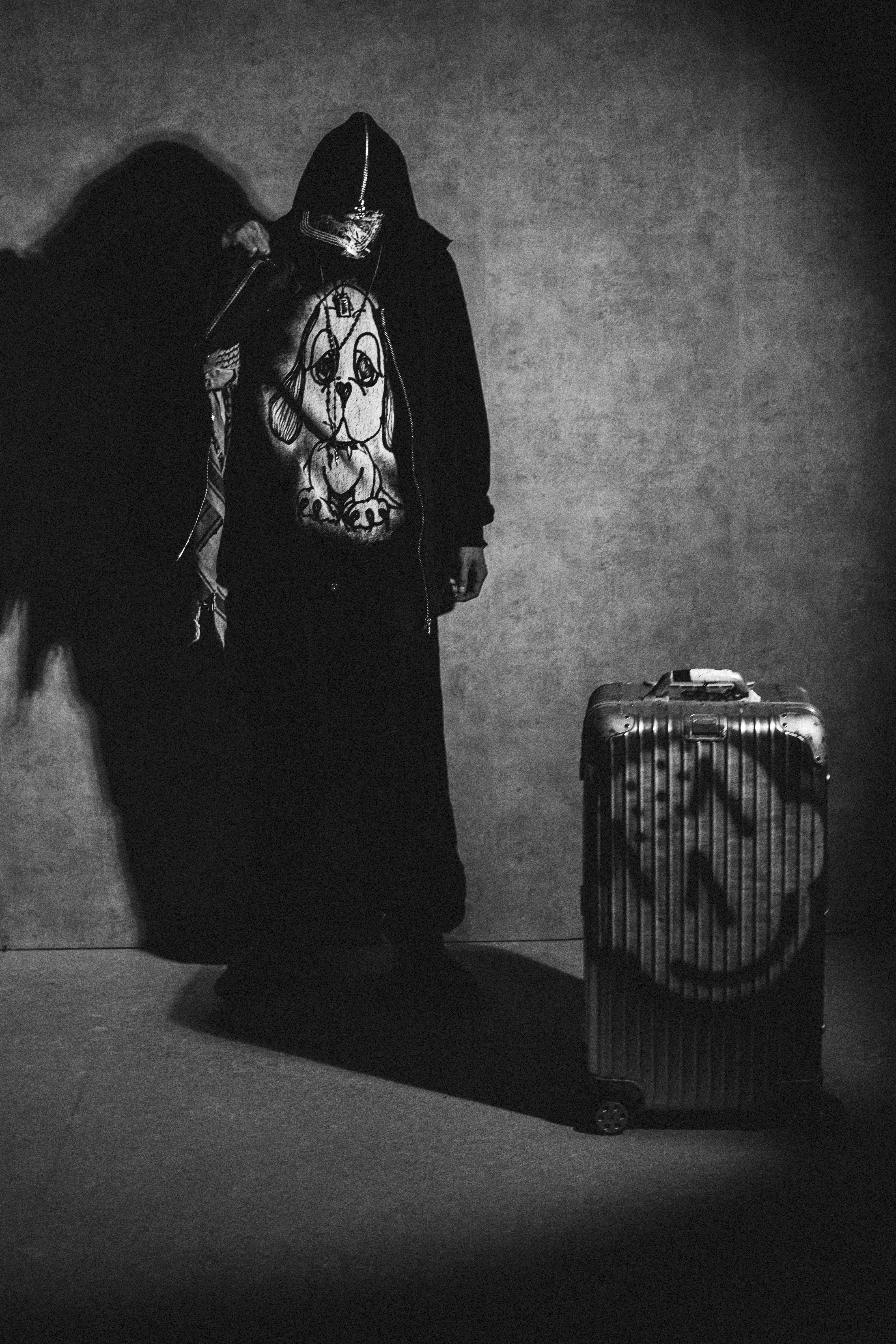A person stands in a dimly lit space, wearing a hoodie and a mask, partially obscured by shadows, next to a suitcase with a peace symbol on it