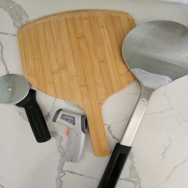 Pizza paddle, pizza cutter, and an infrared thermometer on a marble countertop