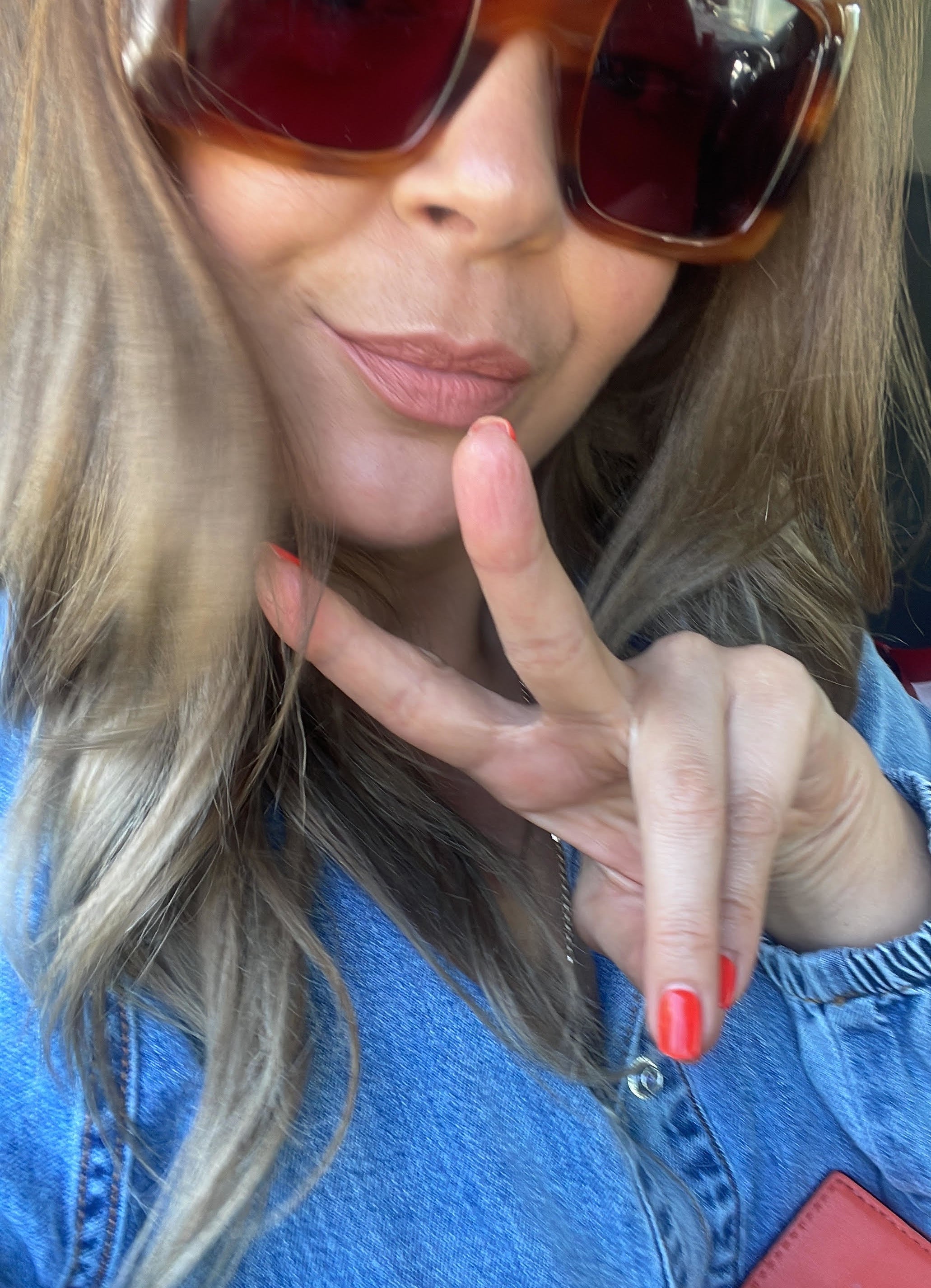 Woman wearing large sunglasses and a denim top shows a peace sign with her fingers while seated in a car