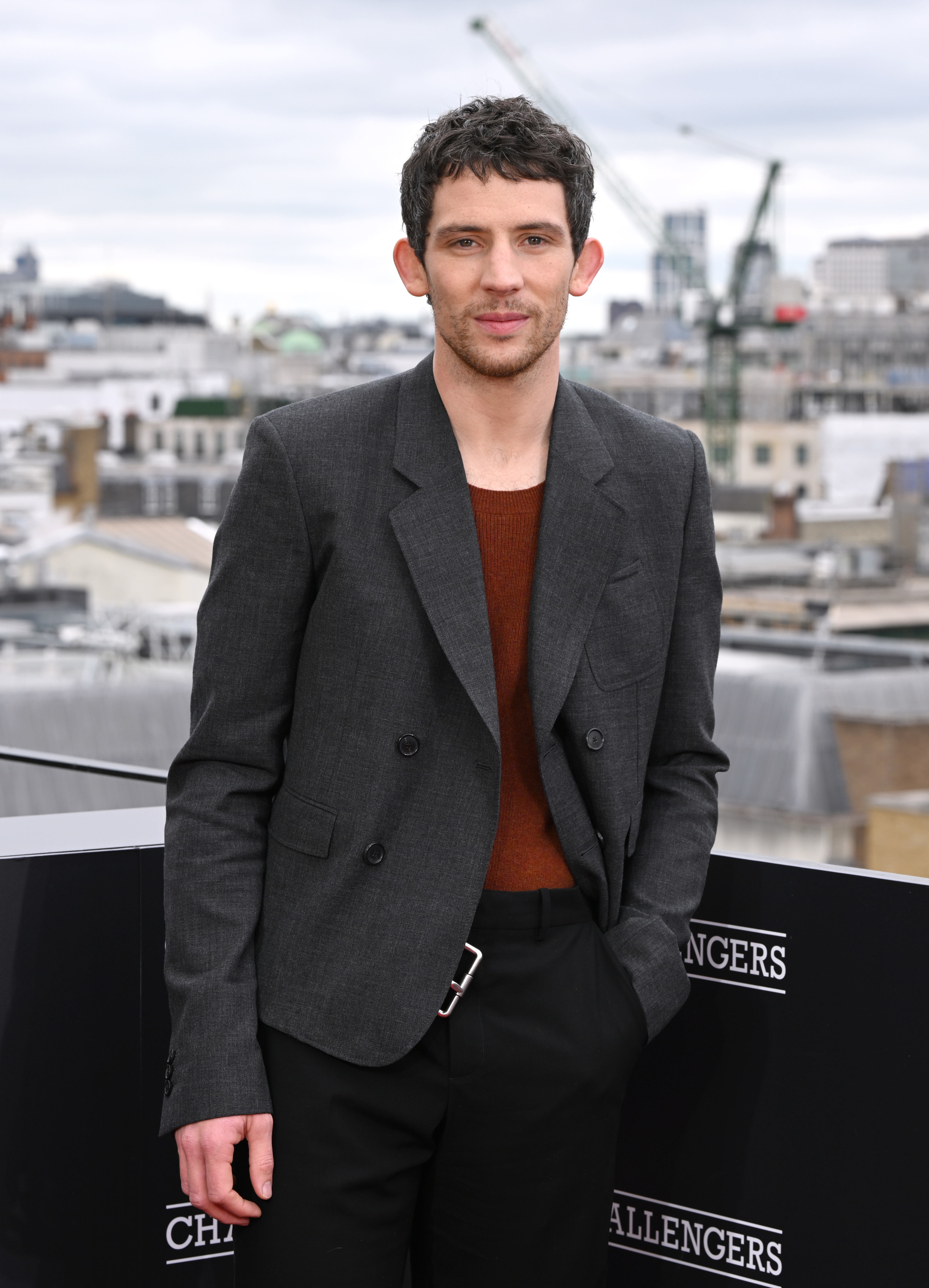 Josh O'Connor stands outdoors, wearing a dark-ish blazer over a maroon shirt, with a cityscape and construction cranes in the background during an event