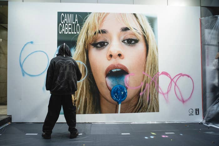 Person in a hoodie spray-painting on a wall-sized image of Camila Cabello holding a blue lollipop in her mouth