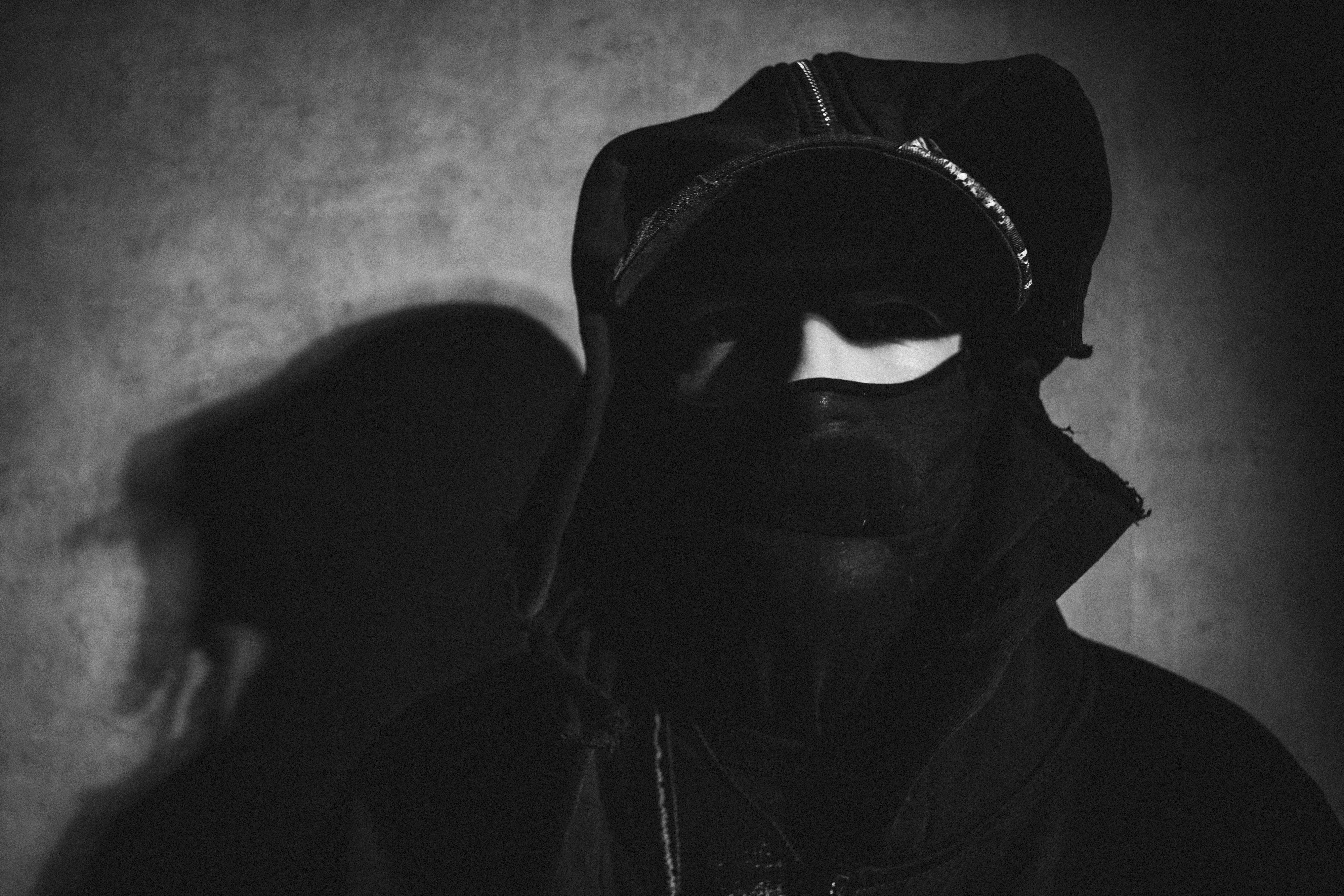 Person wearing a dark, hooded outfit with a mask partially covering their face, creating a mysterious appearance. Background shadow is prominent