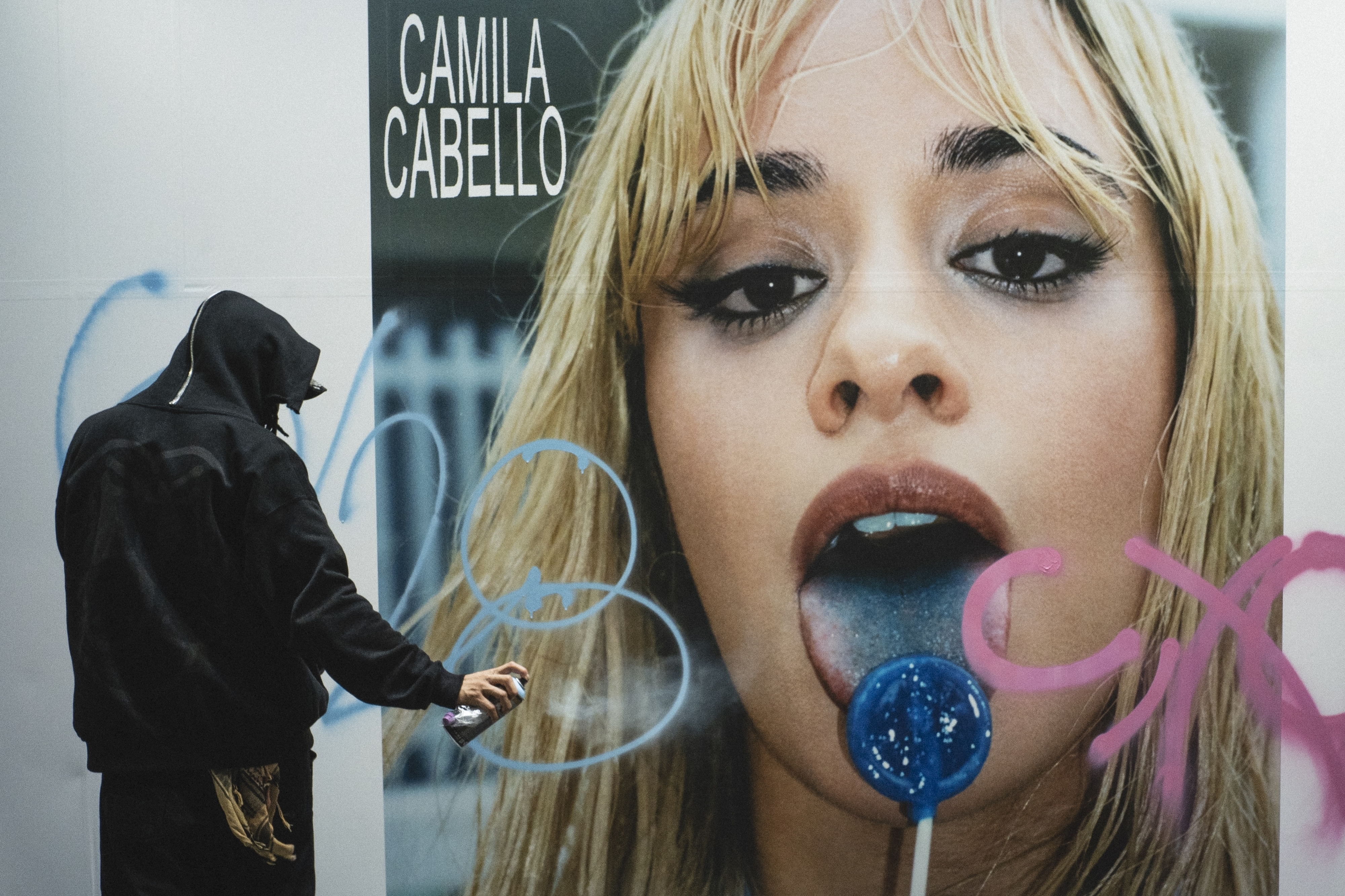 A hooded person spray-paints graffiti on a large poster of Camila Cabello licking a blue lollipop