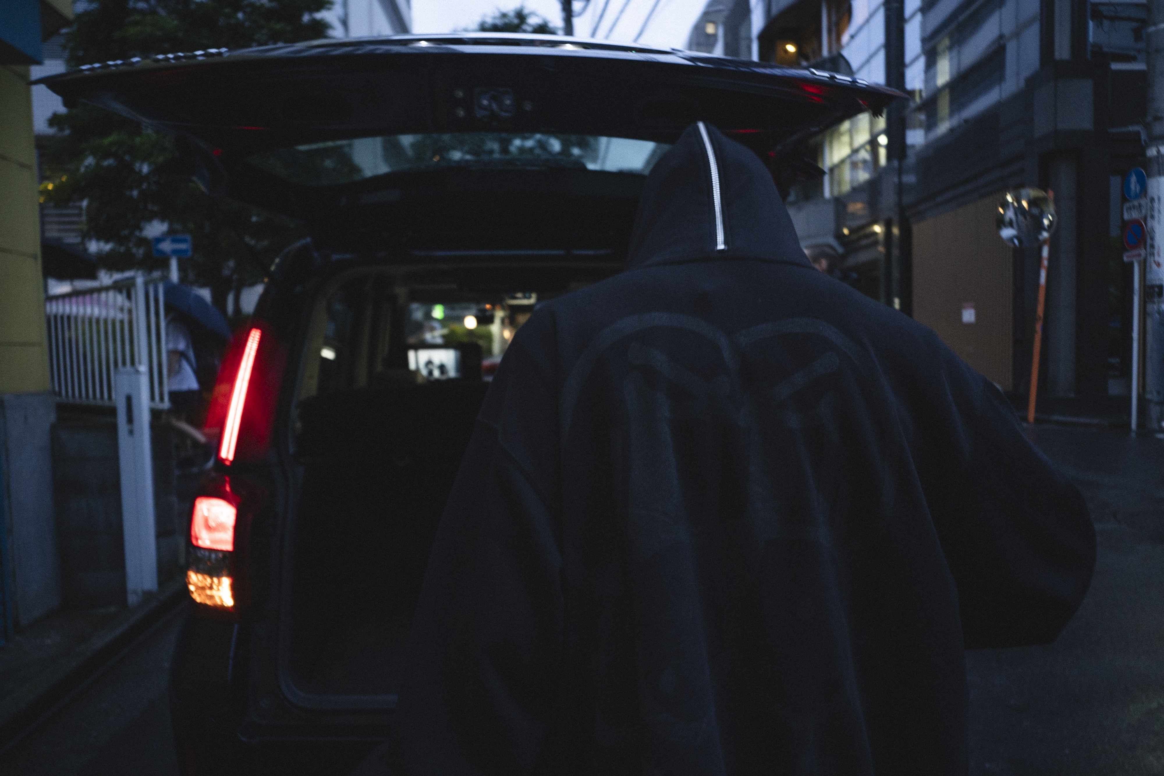 Person in a hooded jacket seen from the back standing near an open car trunk in a dimly lit urban area