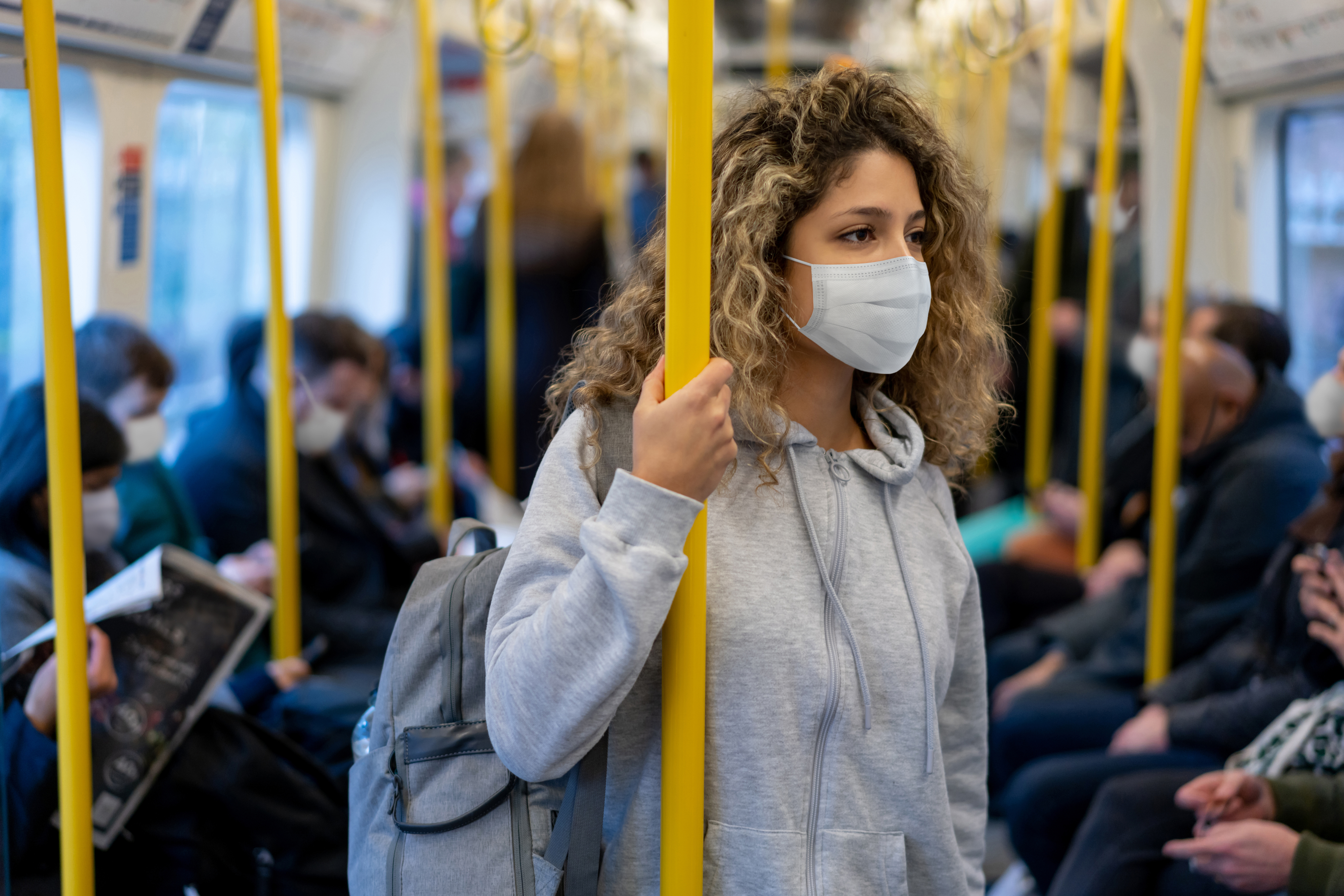 Woman in a gray hoodie and wearing a face mask stands holding a pole on a crowded subway. Other passengers, also masked, are seated and standing around her