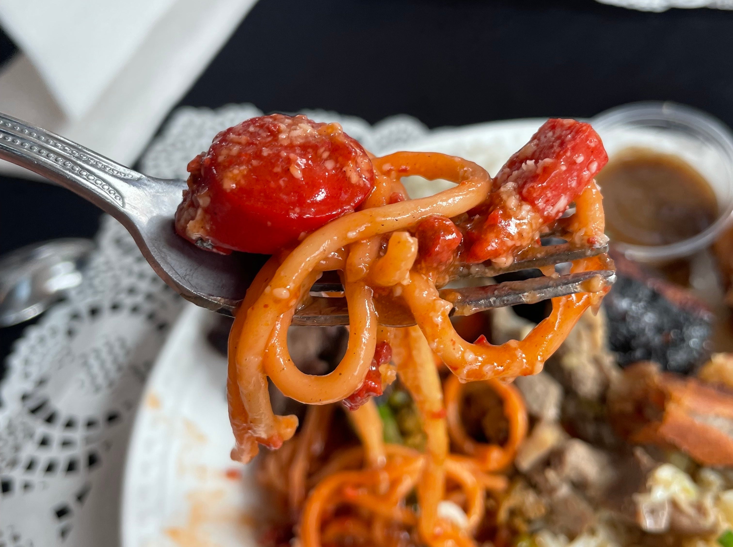 A fork holding a bite of spaghetti with hot dogs s and a white plate with various foods in the background