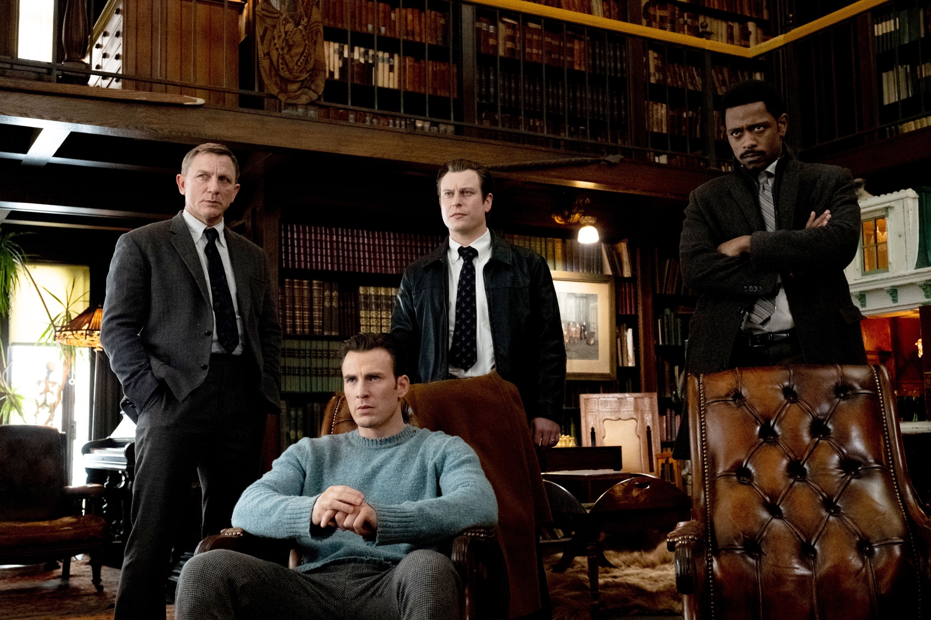 Daniel Craig, Chris Evans, Noah Segan, and Lakeith Stanfield are in a study with bookshelves. Craig and Stanfield stand with serious expressions, while Evans sits