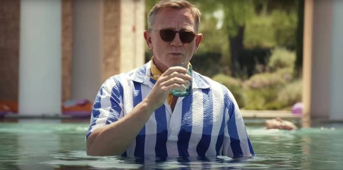 Daniel Craig standing chest-deep in a pool, wearing a striped shirt and sunglasses, holding a drink