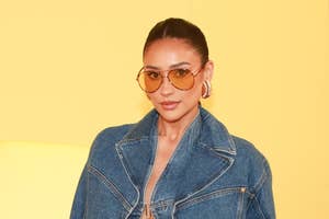 Shay Mitchell wearing a denim jacket and high-waisted jeans, holding a white clutch, and accessorized with large hoop earrings and aviator sunglasses