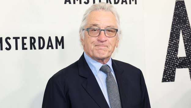 Robert De Niro is standing against a backdrop that reads "Amsterdam," wearing a dark suit, light blue shirt, and gray tie