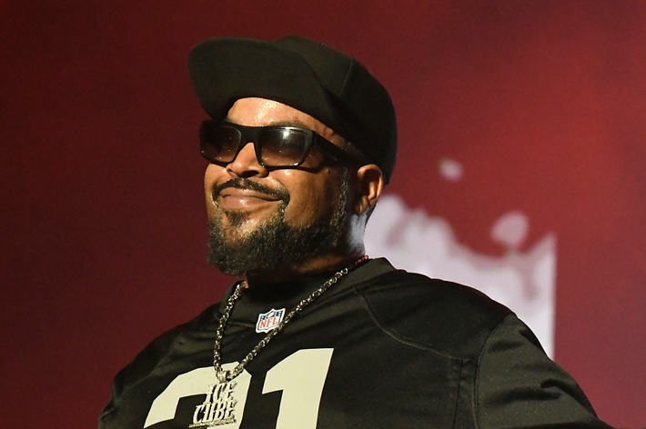 Ice Cube, wearing a black cap, sunglasses, and a black sports jersey with the number 21, performs on stage with a smile
