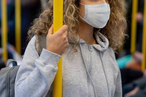 A person wearing a face mask and a gray hoodie holds onto a pole inside a crowded public transport vehicle