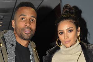 A man wearing casual clothing stands next to Shay Mitchell, who is dressed in camouflage pants, a crop top, a coat, and boots. They pose indoors
