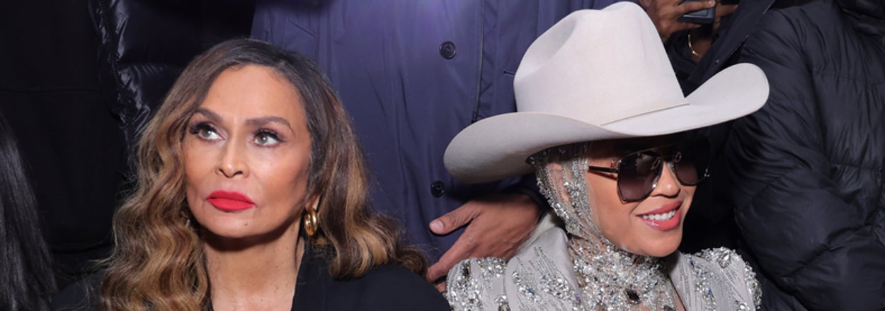 Tina Knowles and Beyoncé attend an event. Tina Knowles wears a black outfit with brown gloves. Beyoncé is in an ornate suit and white hat, holding a holographic bag