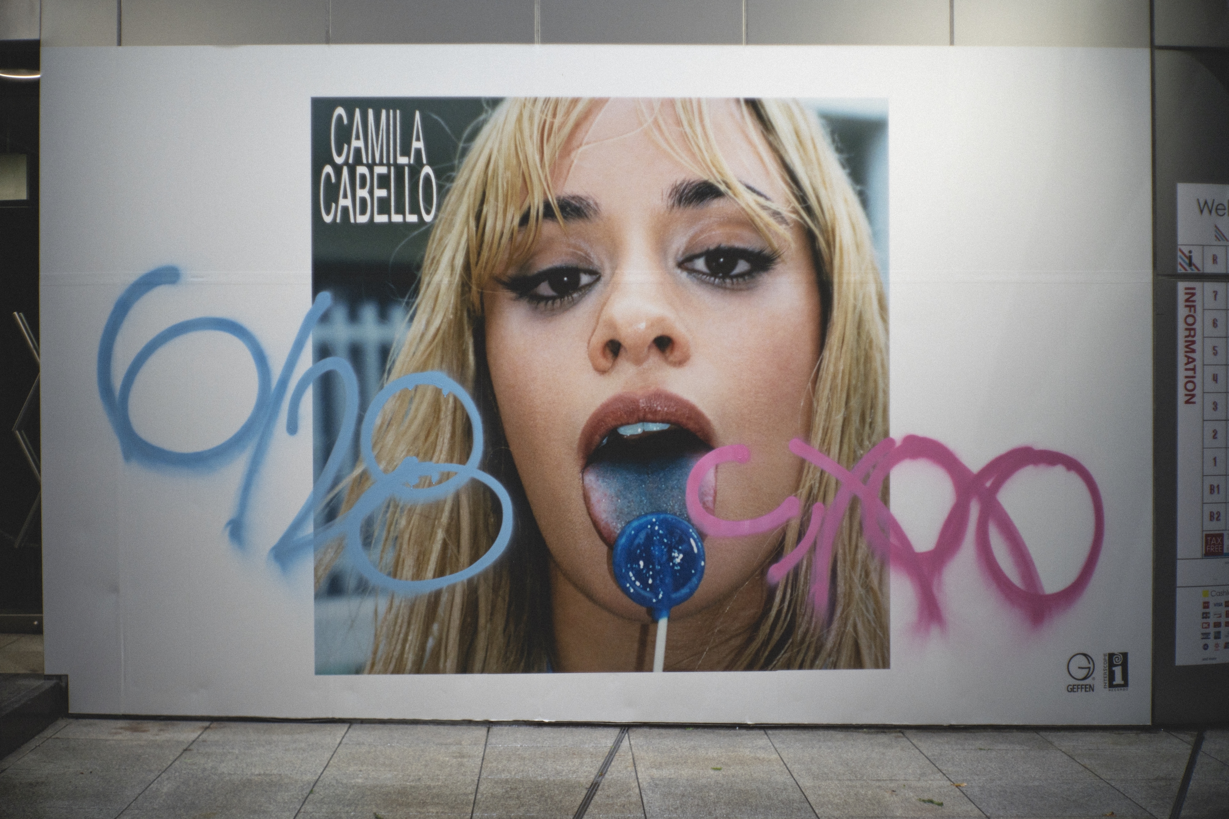 Camila Cabello billboard featuring her licking a blue lollipop, with blue and pink graffiti on the image