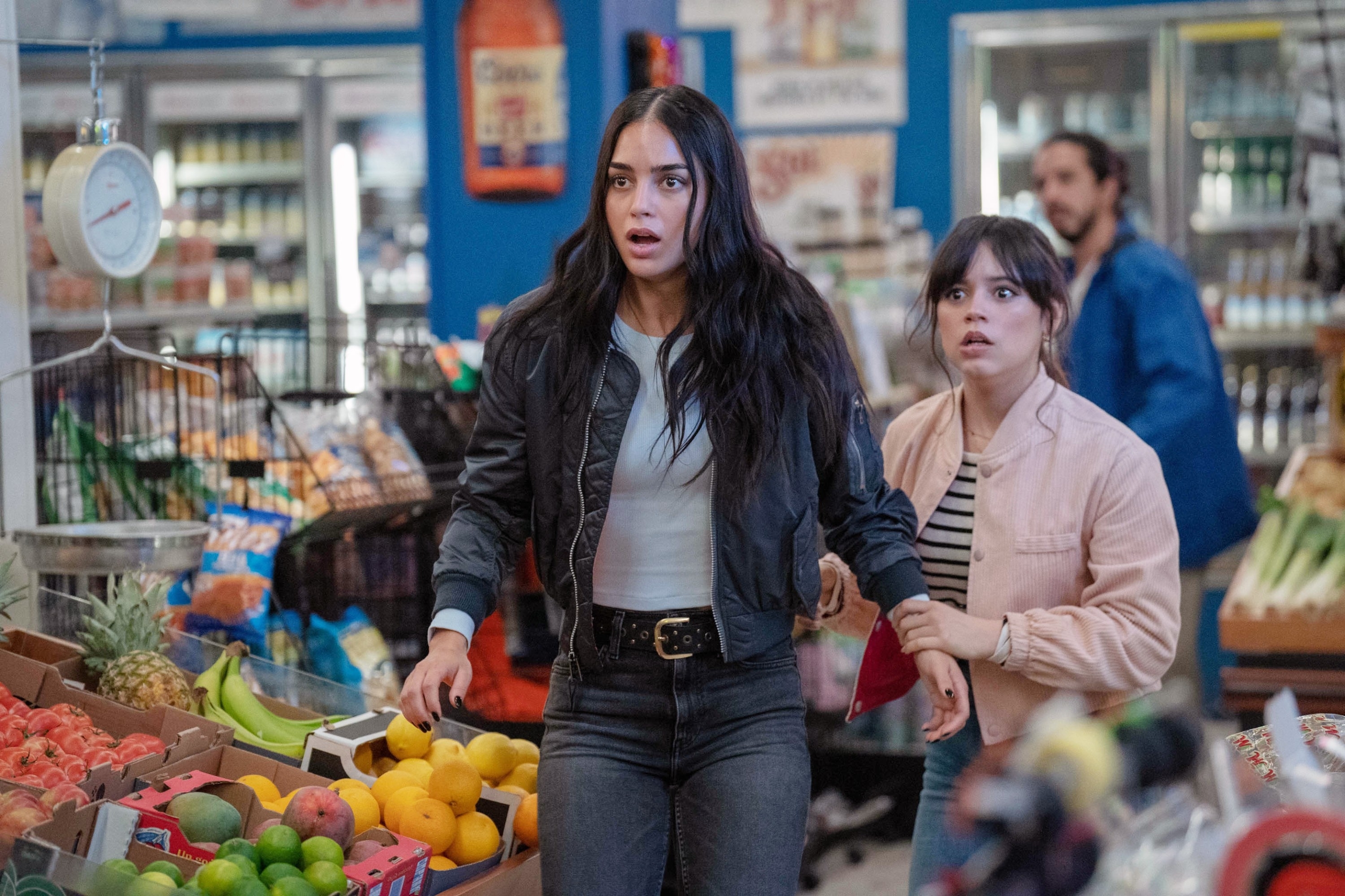 Melissa Barrera and Jenna Ortega in a grocery store, looking surprised. Shelves of various produce can be seen around them