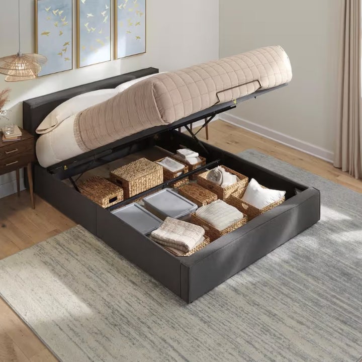 A bed frame with a lift-up mattress reveals organized storage space underneath, featuring baskets and bins containing various household items