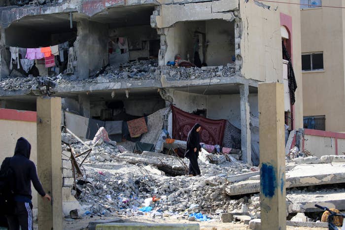 Two people walk through the rubble of a heavily damaged building with exposed rooms and hanging clothes