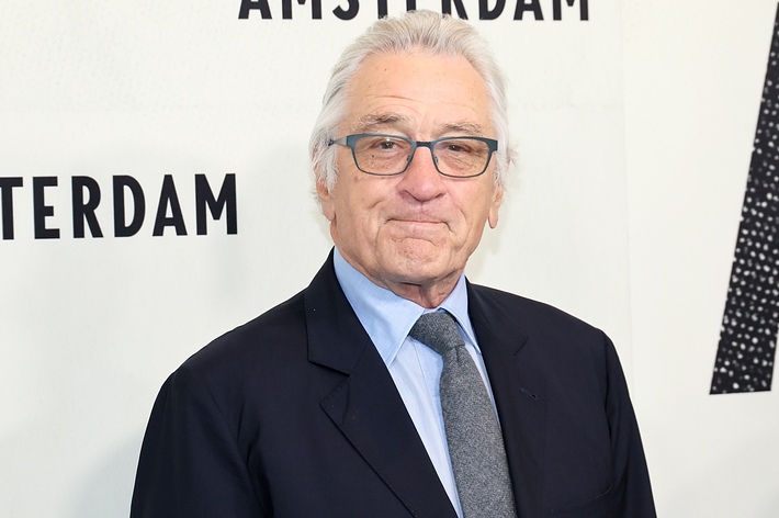 Robert De Niro smiles wearing glasses, a dark suit, light blue shirt, and tie, at the "Amsterdam" event backdrop