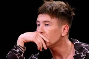 Barry Keoghan is seated, resting his chin on his hand, wearing a printed shirt and a gold chain