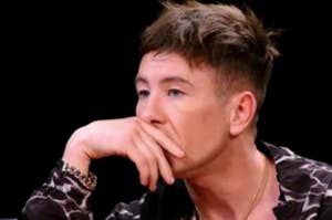 Barry Keoghan is seated, resting his chin on his hand, wearing a printed shirt and a gold chain