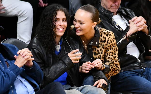 070 Shake and Lily-Rose Depp sit courtside at a sports event. Lourdes wears a black leather jacket, and Lily-Rose sports a leopard print coat