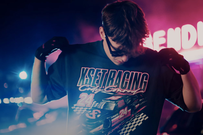 Person showing off a "KSET Racing Cup Series" T-shirt with a race car graphic. The person is wearing sunglasses and gloves, with neon signs in the background
